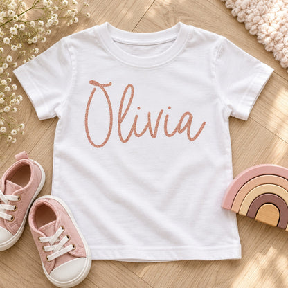 Rose gold glitter iron-on name 'Olivia' on a kids' white T-shirt